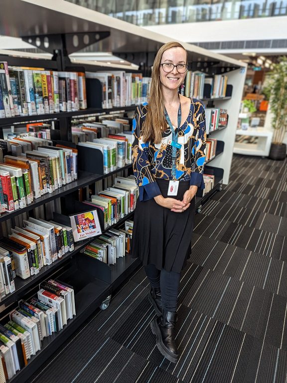 Photo of librarian standing next to library shelves Photo of librarian standing next to library shelves